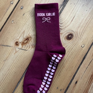 May include: A burgundy sock with the text "BOOK GIRLIE" in white script and a bow graphic. The sole has a line of white heart-shaped grips. The sock is shown on a wooden surface.