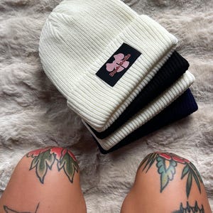 Può includere: Una pila di tre berretti lavorati a maglia in bianco, nero e blu navy. Il berretto bianco in cima ha un'etichetta nera con testo floreale rosa che dice "Book Girlie". I berretti sono impilati su una superficie soffice e di colore chiaro.