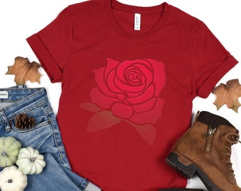 May include: A red t-shirt featuring a large, detailed rose graphic. The shirt is paired with blue denim jeans, brown leather boots, and decorative fall elements. The t-shirt is made of a soft material.