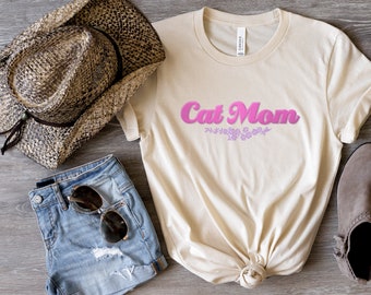 Könnte beinhalten: Ein beiges T-Shirt mit dem Text "Cat Mom" in rosa Buchstaben mit einem floralen Design darunter. Das Shirt liegt auf einer Holzoberfläche mit einem Strohhut, Jeans-Shorts, Sonnenbrille und einem braunen Schuh.