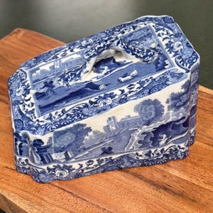 May include: Blue and white ceramic butter dish with a handle. The dish features a landscape design with trees, buildings, and figures. The edges are decorated with floral patterns. The dish is rectangular with a slightly angled top.