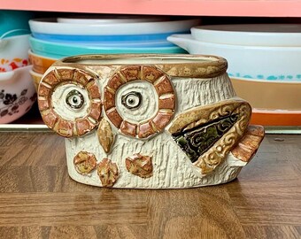 May include: A ceramic owl-shaped planter with brown and beige tones. The owl has large, round eyes and is perched on a wooden surface. A collection of vintage Pyrex bowls in various colors and patterns are visible on shelves in the background.