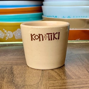 May include: A tan ceramic cup with the word "Kon Tiki" in brown lettering. The cup is cylindrical and sits on a wooden surface. Various vintage bowls and dishes are visible in the background.
