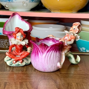 May include: Two vintage ceramic figurines of fairies sitting in flower-shaped planters. The fairy on the left is wearing a red dress and a white collar. The fairy on the right is wearing a pink dress and a white collar. Both fairies have pink wings and are sitting in flower-shaped planters.