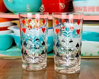 May include: Two clear glass drinking glasses with a white floral design and red and black playing card symbols. The glasses are on a wooden surface.