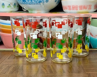 May include: Set of six vintage clear drinking glasses. Each glass features a repeating pattern of red, white, green, and yellow squares. The glasses are displayed on a wooden surface, with shelves of vintage kitchenware in the background.