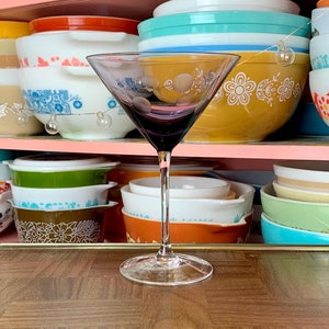 May include: A clear glass martini glass with a purple tint sits on a brown wooden table. The glass is empty and has a stem and a wide bowl. The table has a checkered pattern.