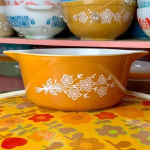 May include: A yellow ceramic casserole dish with a white floral design. The dish is sitting on a yellow and orange floral patterned serving tray.
