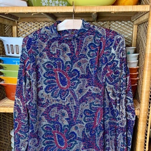 May include: A blue and red patterned long-sleeved shirt with a high neck, hanging on a wooden rack with a variety of colorful bowls stacked on shelves.