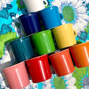 May include: A set of ten colorful ceramic mugs, arranged in a pyramid shape. The mugs are in various shades of blue, green, yellow, red, and pink. The mugs are sitting on a blue and white floral tablecloth.