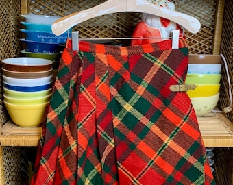 May include: A red, green, and yellow plaid skirt with a brown leather belt buckle. The skirt is hanging on a white wooden hanger in front of a wicker shelf with bowls and cups.