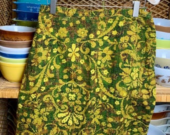 May include: A green and yellow floral print skirt hanging on a white wooden hanger. The skirt is made of a textured fabric and has a vintage style.
