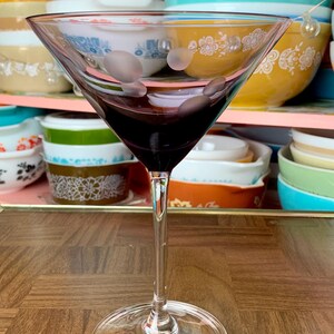 May include: A clear glass martini glass with a frosted design and a dark purple liquid inside. The glass is sitting on a wooden table.