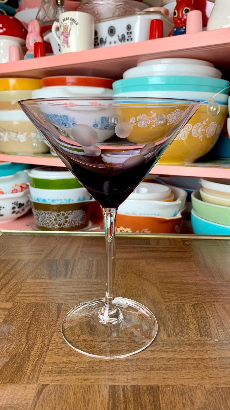 May include: A clear glass martini glass with a frosted design and a dark purple liquid inside. The glass is sitting on a wooden table.