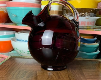 May include: A dark purple glass pitcher with a clear handle. The pitcher is sitting on a wooden surface.