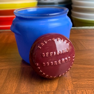 May include: A blue ceramic jar with a burgundy lid. The lid has the words "Tri-State Official Door Knob Pottery" embossed on it.