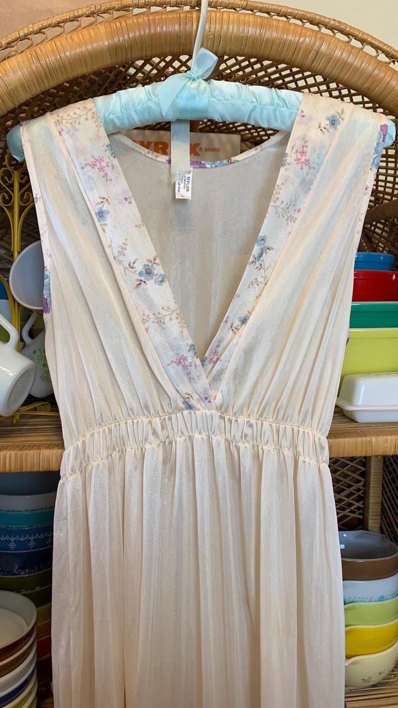 May include: A white sleeveless nightgown with a floral print and a V-neckline. The neckline is trimmed with a floral lace. The nightgown is made of a sheer fabric.