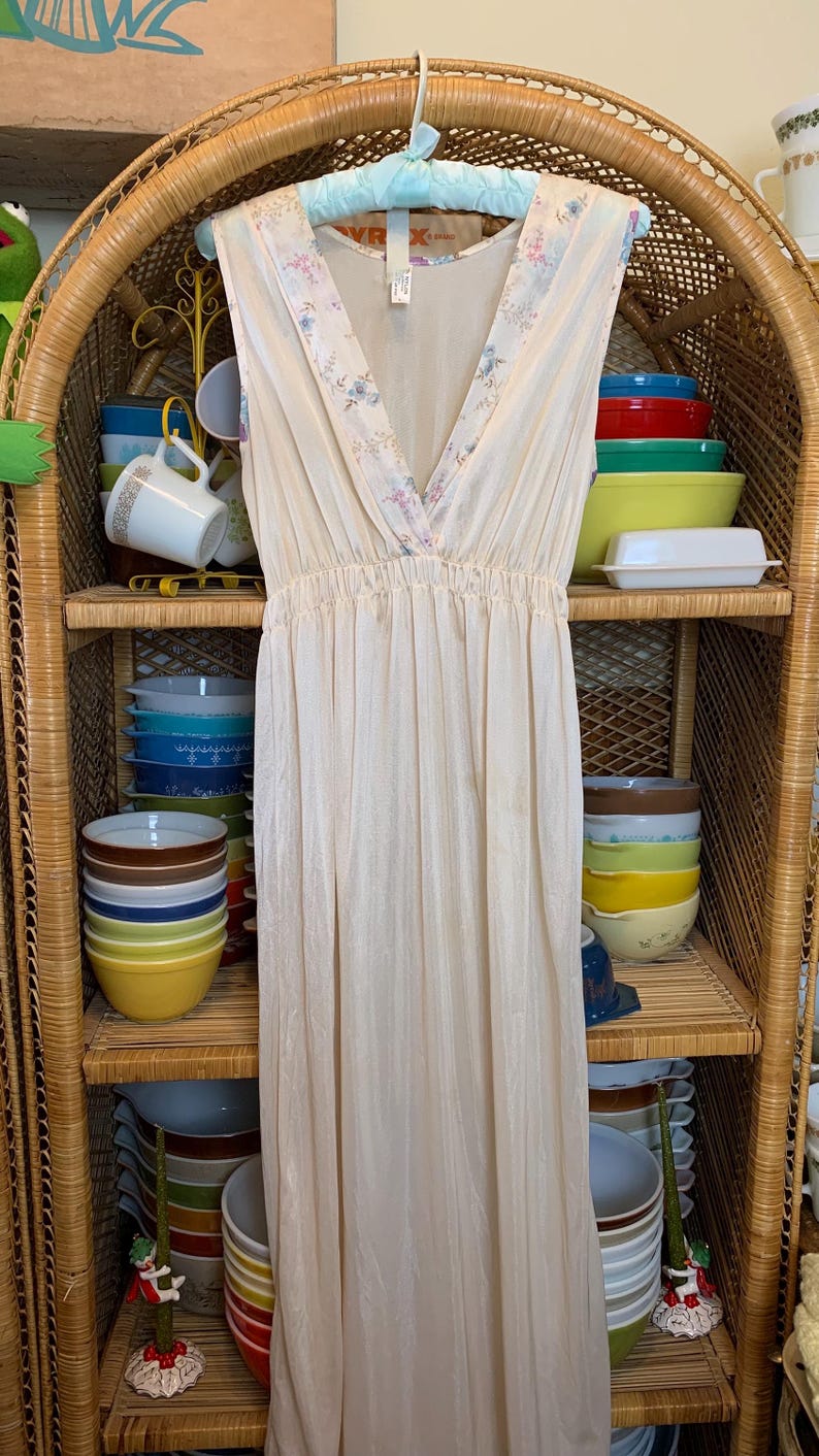 May include: A white sleeveless maxi dress with a floral print on the bodice. The dress is hanging on a blue hanger in front of a wicker shelf with bowls.