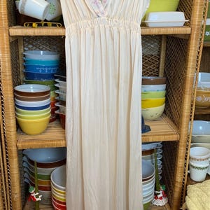 May include: A white sleeveless nightgown with a floral print trim around the neckline. The nightgown is hanging on a wicker shelf with colorful bowls stacked on the shelves.