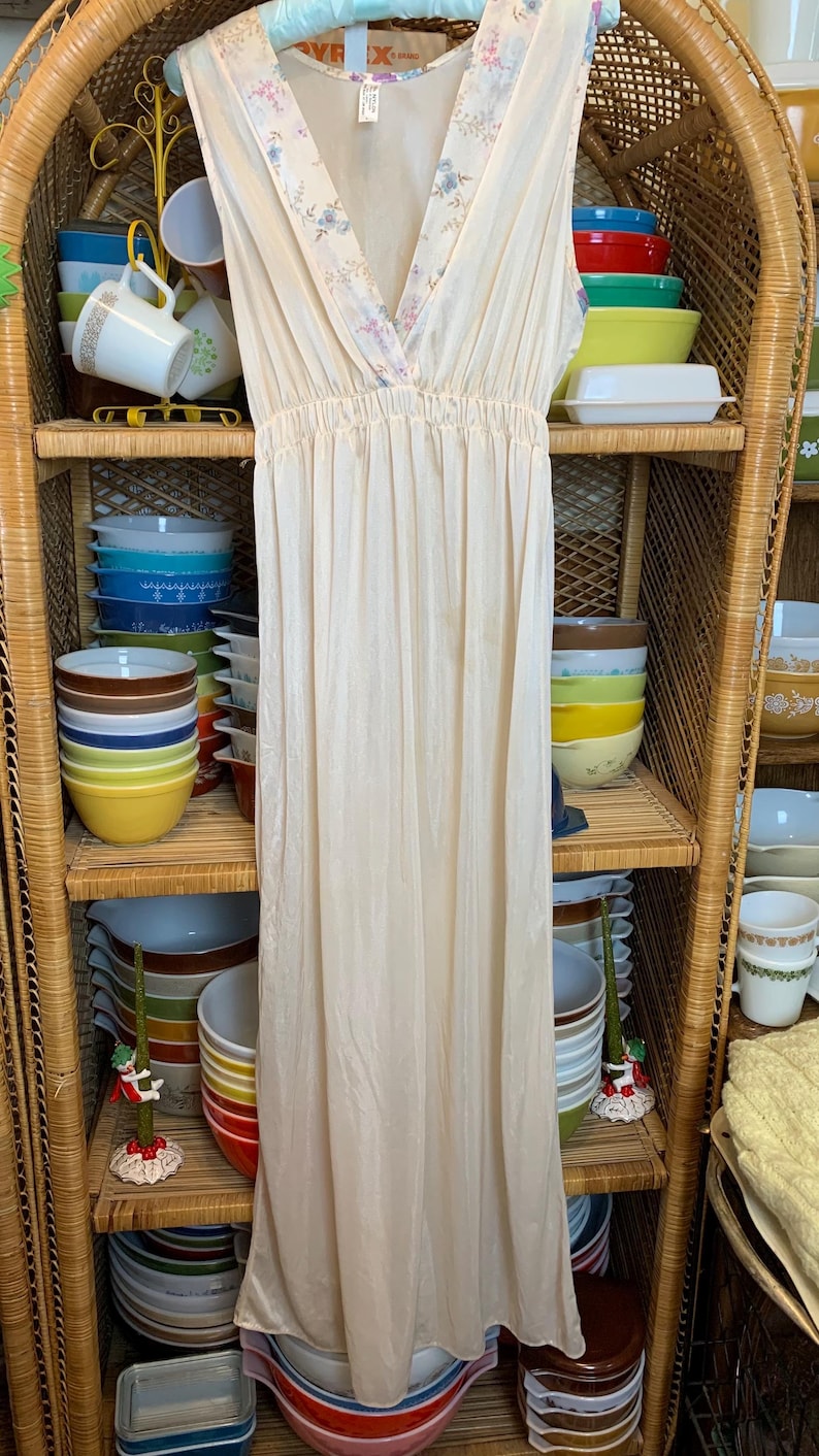 May include: A white sleeveless nightgown with a floral print trim around the neckline. The nightgown is hanging on a wicker shelf with colorful bowls stacked on the shelves.