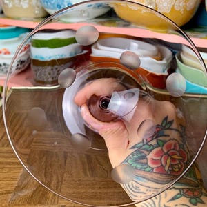 May include: A clear glass cake stand with a raised edge and a pattern of small, raised dots on the bottom. The stand is being held by a hand with a tattoo.