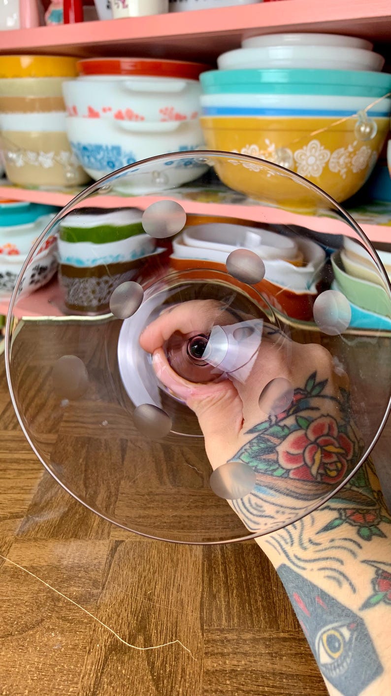 May include: A clear glass cake stand with a raised edge and a pattern of small, raised dots on the bottom. The stand is being held by a hand with a tattoo.