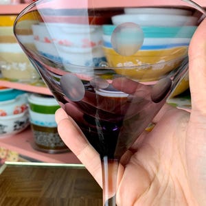 May include: A clear glass martini glass with a purple tint and white polka dots. The glass is being held by a hand.