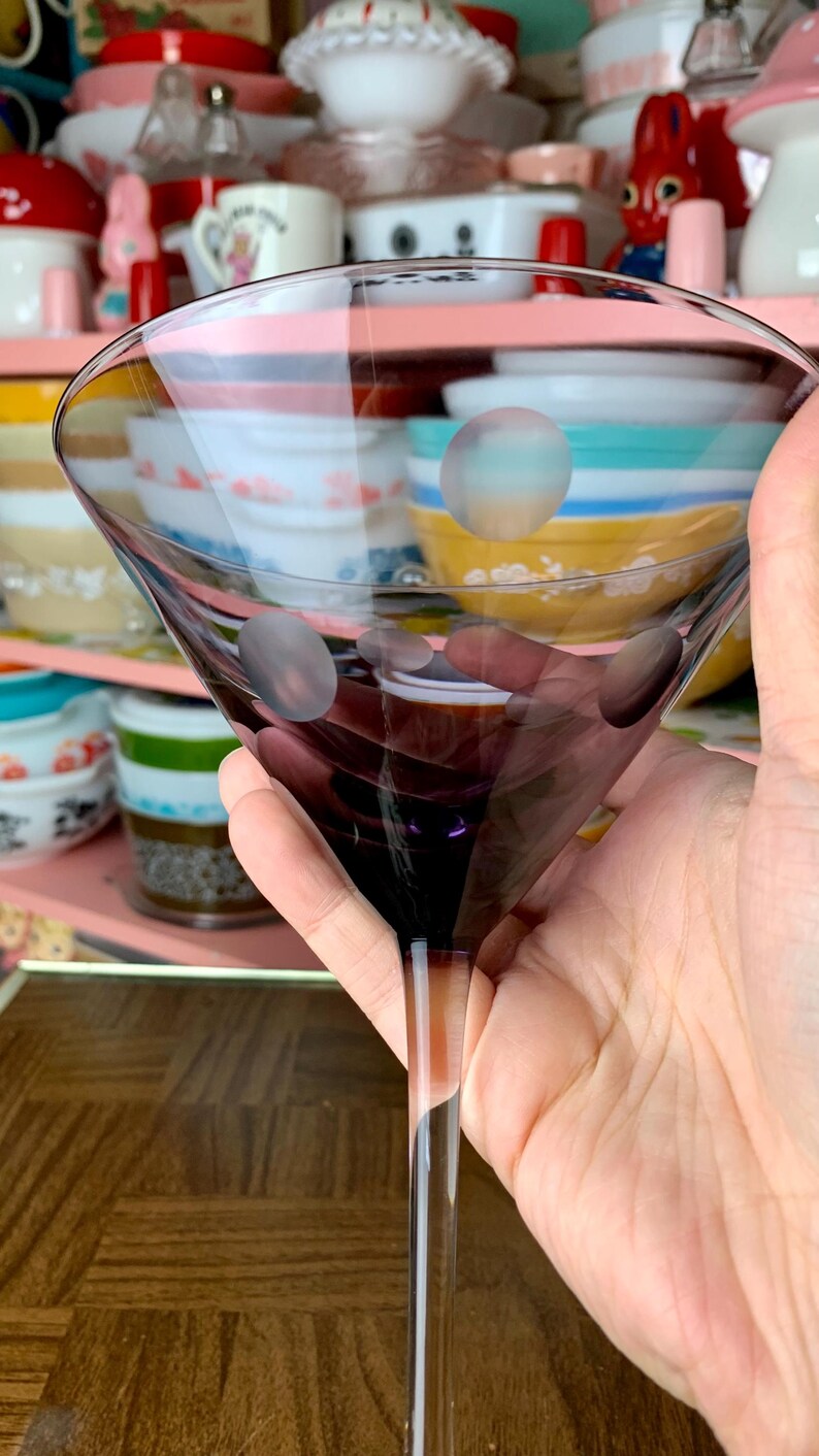 May include: A clear glass martini glass with a purple tint and white polka dots. The glass is being held by a hand.