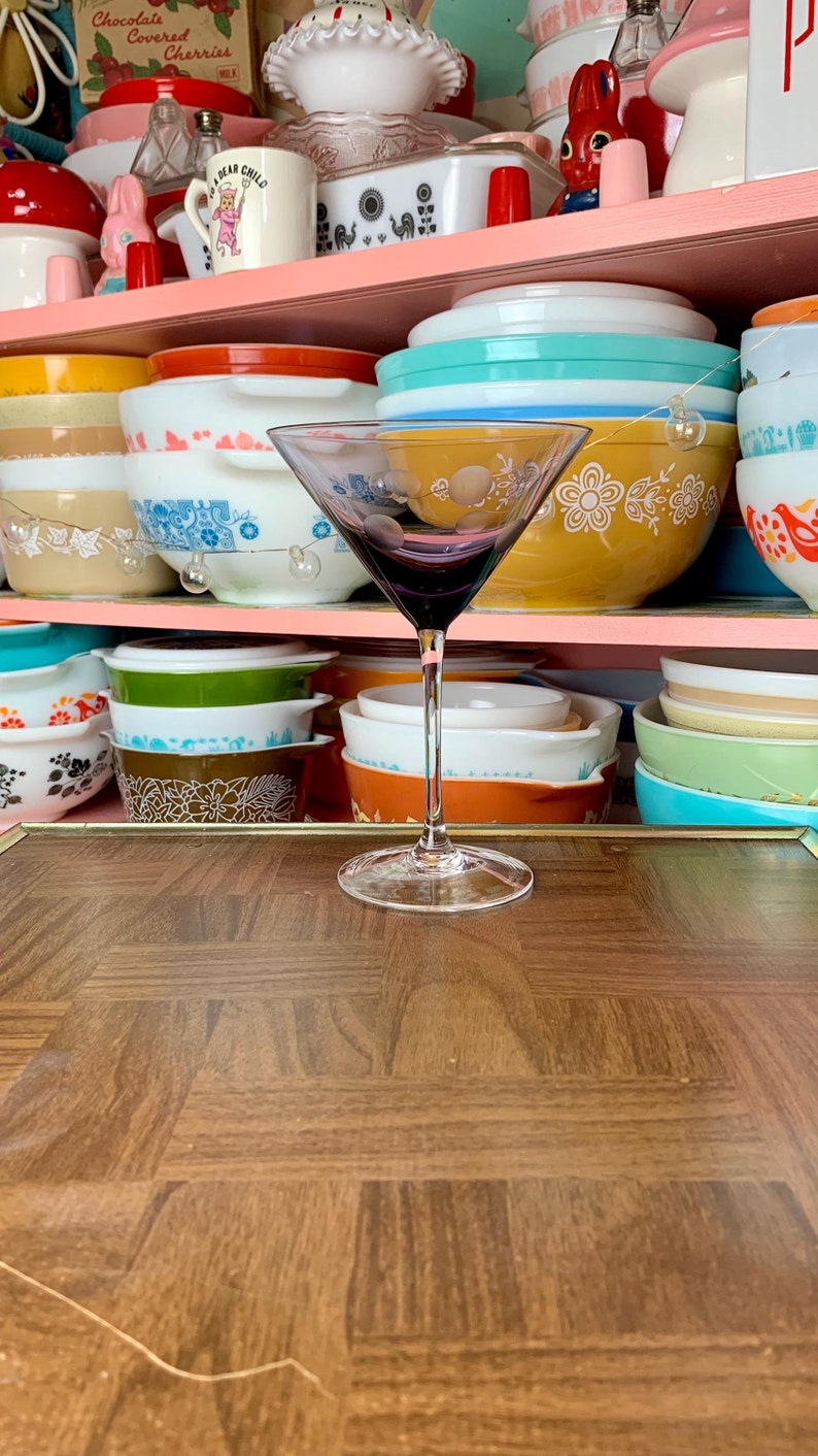 May include: A clear glass martini glass with a purple tint sits on a brown wooden table. The glass is empty and has a stem and a wide bowl. The table has a checkered pattern.