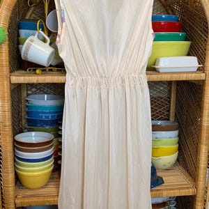 May include: A long, sleeveless, white slip dress with a gathered waistline. The dress is hanging on a hanger in front of a wicker shelf with bowls and mugs.