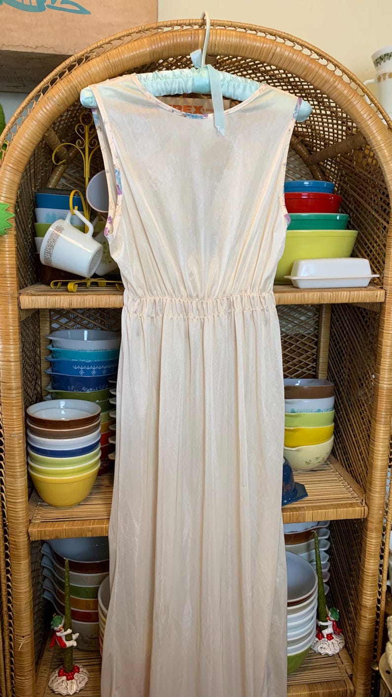 May include: A long, sleeveless, white slip dress with a gathered waistline. The dress is hanging on a hanger in front of a wicker shelf with bowls and mugs.