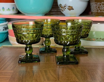 May include: A collection of vintage green glass goblets with diamond-patterned bowls and square bases. The goblets are displayed on a wooden surface, with a backdrop of colorful vintage bowls and kitchenware.