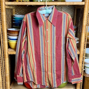 May include: A long-sleeved button-down shirt with a red, yellow, and blue striped pattern. The shirt has a collared neckline and a single chest pocket. The shirt is hanging on a wooden shelf with various bowls and cups.