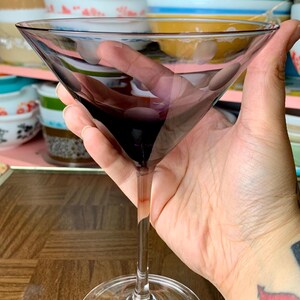 May include: A clear glass martini glass with a purple tint. The glass is held in a hand with a tattoo.