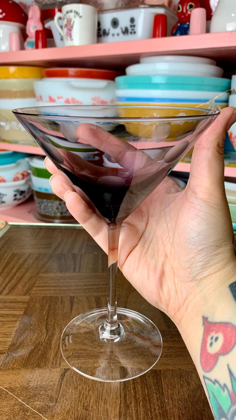 May include: A clear glass martini glass with a purple tint. The glass is held in a hand with a tattoo.