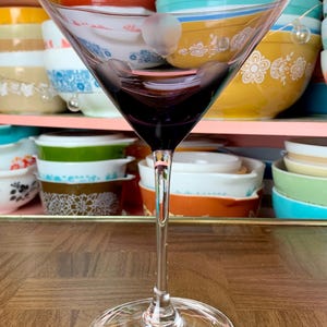 May include: A clear glass martini glass with a purple hue sits on a wooden surface. The glass is empty and appears to be in good condition.