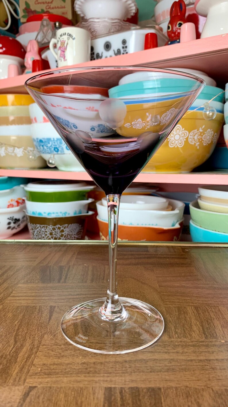 May include: A clear glass martini glass with a purple hue sits on a wooden surface. The glass is empty and appears to be in good condition.