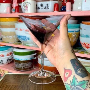 May include: A clear glass martini glass with a purple liquid inside. The glass is being held by a hand with a tattoo on the forearm.
