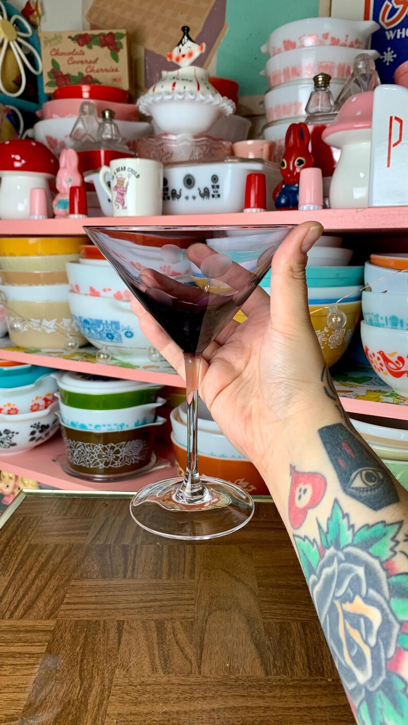 May include: A clear glass martini glass with a purple liquid inside. The glass is being held by a hand with a tattoo on the forearm.