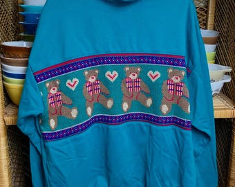 May include: Teal blue sweatshirt with a festive pattern of brown teddy bears holding red and white striped gifts. The bears are surrounded by a green, purple, and red patterned border.