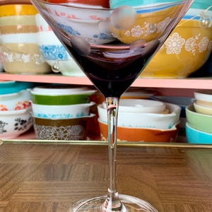May include: A clear glass martini glass with a purple tint sits on a wooden surface. The glass is empty and has a long stem.