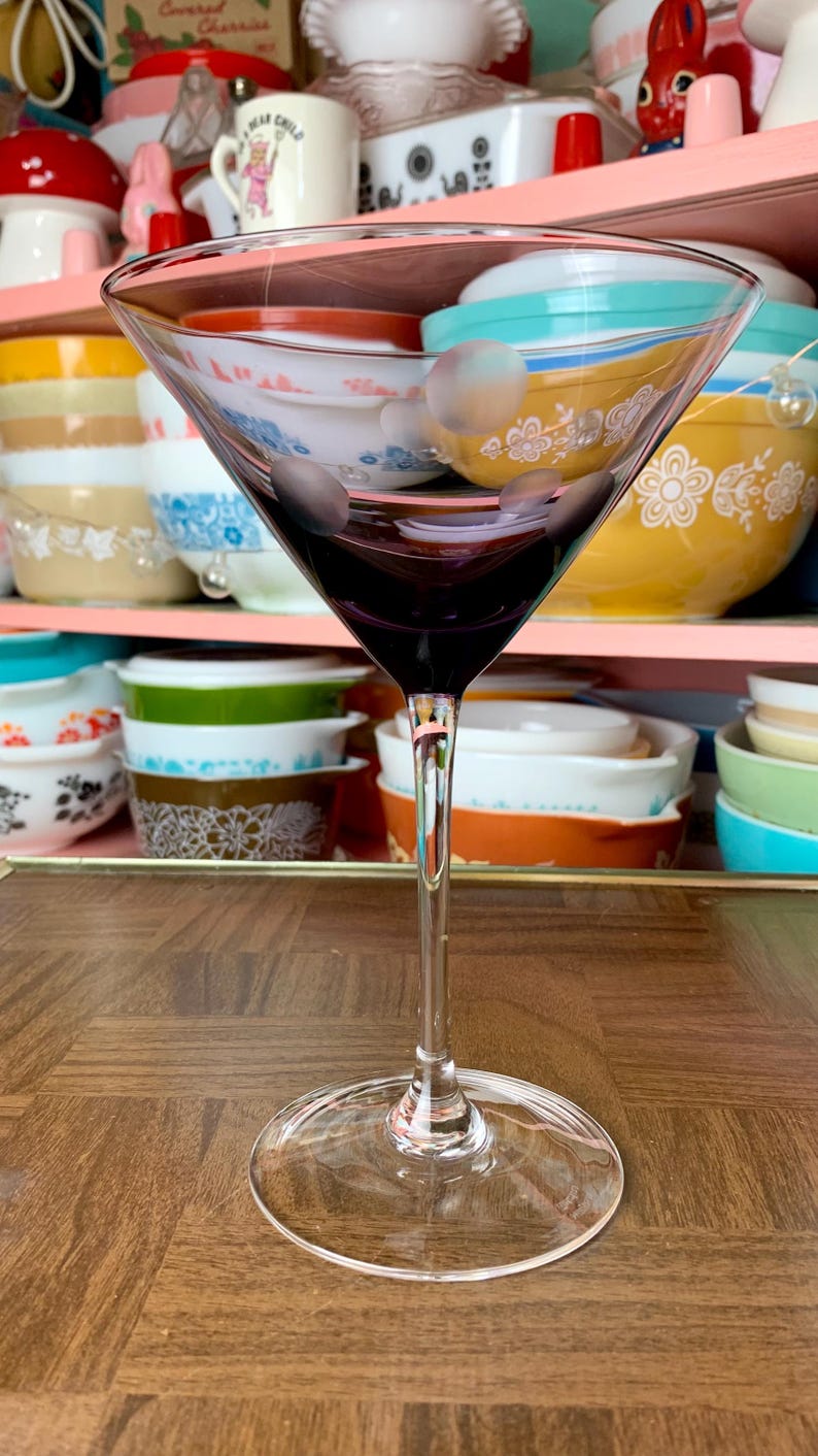 May include: A clear glass martini glass with a purple tint sits on a wooden surface. The glass is empty and has a long stem.