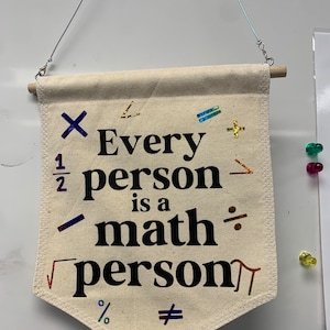 May include: A beige canvas banner with the text "Every person is a math person" in black. The banner includes various math symbols in a mix of colors. It is suspended by a wooden dowel and wire.