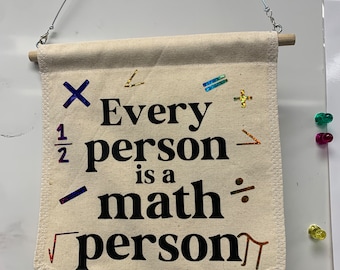 May include: A beige canvas banner with the text "Every person is a math person" in black. The banner includes various math symbols in a mix of colors. It is suspended by a wooden dowel and wire.