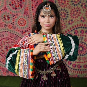 Könnte beinhalten: Eine Frau in einem traditionellen afghanischen Kleid mit aufwendigen Stickereien und Verzierungen. Das Kleid hat leuchtende Farben, darunter Rot, Grün und Gold, mit einem schwarzen Rock. Sie ist mit Kopfschmuck und Schmuck geschmückt und präsentiert kulturelle Kleidung.