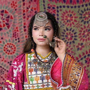 May include: A woman wearing a traditional pink dress with intricate embroidery and embellishments. The dress features a detailed neckline and sleeves, complemented by silver jewelry, including a headpiece and nose ring. The background showcases patterned textiles.