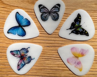 Könnte beinhalten: Fünf weiße Gitarrenplektren mit Schmetterlingsmotiven. Die Schmetterlinge weisen verschiedene Farben auf, darunter Blau, Rosa und Schwarz, mit detaillierten Flügelmustern. Die Plektren liegen auf einer Holzoberfläche.
