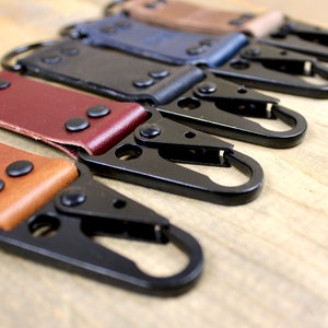 May include: Five leather keychains with black metal clips. The keychains are in different colors: brown, burgundy, black, navy blue, and tan. The keychains have a metal ring at the top and a metal clip at the bottom. The keychains are arranged in a row on a wooden surface.