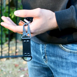 May include: A black leather key chain with a metal clip and a ring. The key chain is attached to a pair of keys.