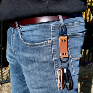 May include: A brown leather key chain with a black metal clip and two keys attached. The key chain is attached to a belt loop on a pair of blue jeans.